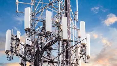 Close-up of a cell tower with multiple white antennas and black cables, set against a blue, cloudy sky.