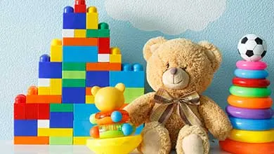 Children’s toys: a brown teddy bear with a plaid bow, a colorful block structure, and a stacking ring toy with a soccer ball on top.