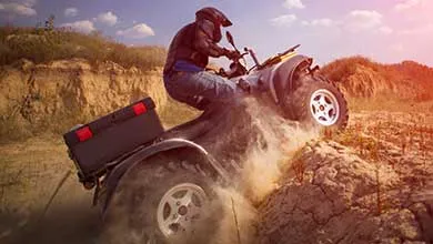 A person in a helmet and jacket rides an ATV up a steep sandy hill, creating a large dust cloud.