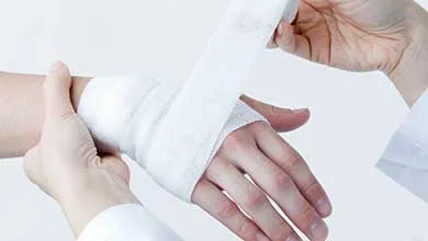 Close-up of one person wrapping another's wrist and hand with a white gauze bandage on a plain white background.