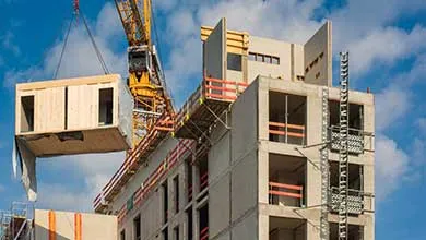 A construction crane lifting a prefabricated concrete module onto a multi-story building under construction against a blue sky.