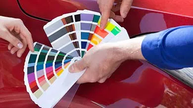 Two hands holding a fan of color swatches against a red car exterior to compare and select matching colors.