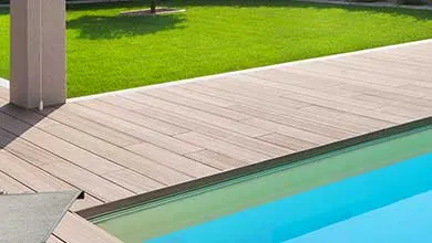 A light brown composite deck borders a bright blue swimming pool, with a green lawn in the background.