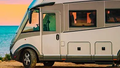 A white and grey RV is parked on a sandy beach by the ocean at sunset.