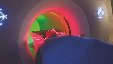 A person lies on a table entering a CT scanner, which is illuminated with red and green lights inside.