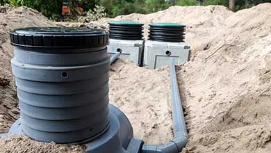 A large, round gray septic tank in the foreground is connected by PVC pipes to two smaller, partially buried tanks in the sandy ground behind it.