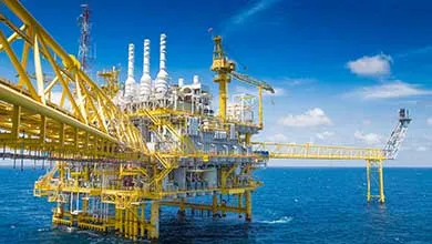 A large yellow and white offshore oil rig stands in the blue ocean beneath a clear sky.